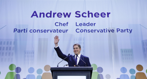 Konservative-Parteichef Andrew Scheer steht an einem Podium mit zwei Mikrofonen und einem Bildschirm dahinter, mit einem Banner mit Text und Gebäudeillustrationen im Hintergrund.