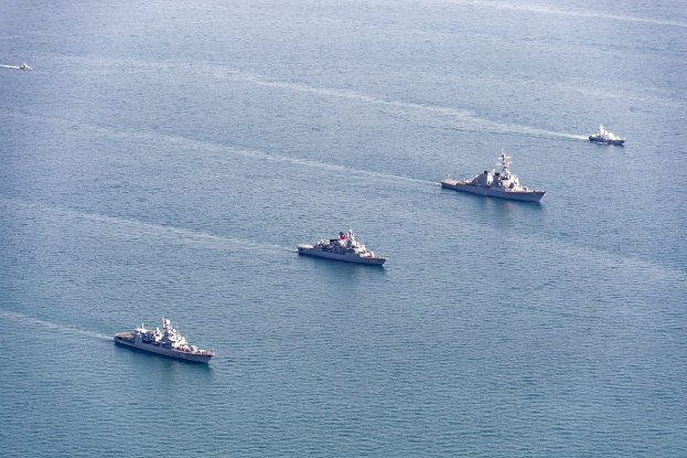 Eine Gruppe von Schiffen verschiedener Größen und Farben, die in tiefblauen Ozeanwassern unter strahlendem Sonnenlicht segeln, mit Menschen an Bord.