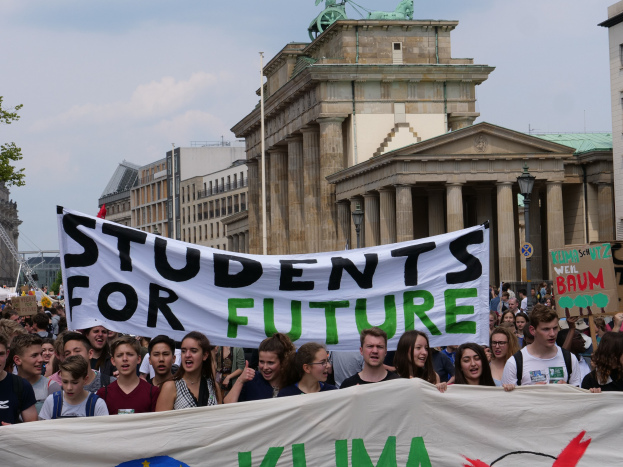 Eine Gruppe von Studenten marschiert in Berlin und hält ein buntes Banner mit der Aufschrift "Students for Future". Im Hintergrund sind Gebäude, Bäume und Himmel zu sehen.