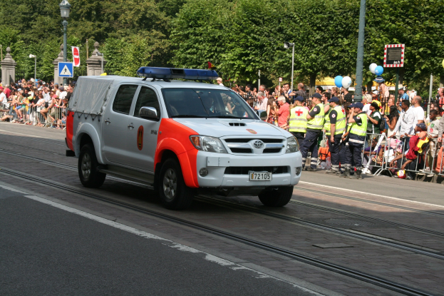 Polizeiauto fährt an einer Menschenmenge auf einer Straße mit Geländern, Laternen, Schildern, Ballons und Bäumen im Hintergrund vorbei.