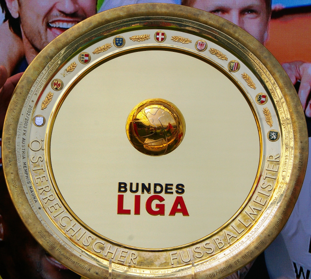 Ein Mann hält einen goldenen Schild mit der Aufschrift "Bundesliga" hoch, im Hintergrund ein Banner mit drei Personen