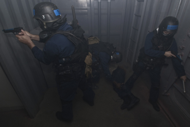 Eine Gruppe von Polizisten in Einsatzausrüstung, einer hält eine Waffe, steht in der Nähe eines Metallbehälters und einer Leiter in einem begrenzten Raum.