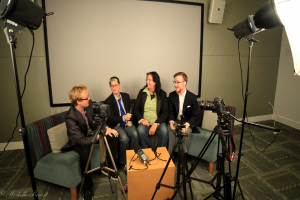 Eine Gruppe von drei Personen sitzt auf einem Sofa und gibt ein Interview, wobei eine Person links in einem schwarzen Mantel und Hose, eine Person in der Mitte in einem Mantel und blauer Krawatte mit einem Mikrofon und eine Person rechts mit einem Lächeln zu sehen ist. Ein Mikrofonrekorder befindet sich auf dem Mitteltisch, Kameras sind auf der rechten und linken Seite und große Scheinwerfer sind vorhanden, mit einem Projektionsschirm im Hintergrund.