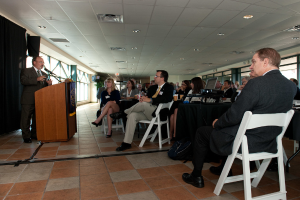 Eine Gruppe von Menschen, darunter Männer und Frauen, sitzt einem Mann gegenüber, der an einem Podium steht und in ein Mikrofon spricht, mit Fenstern im Hintergrund.