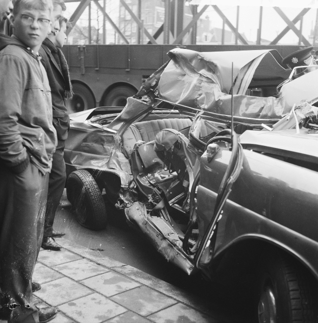 Schwarz-weißes Bild eines verbeulten Autos am Straßenrand mit Menschen in der Nähe, einem Lastwagen und einer Brücke im Hintergrund.