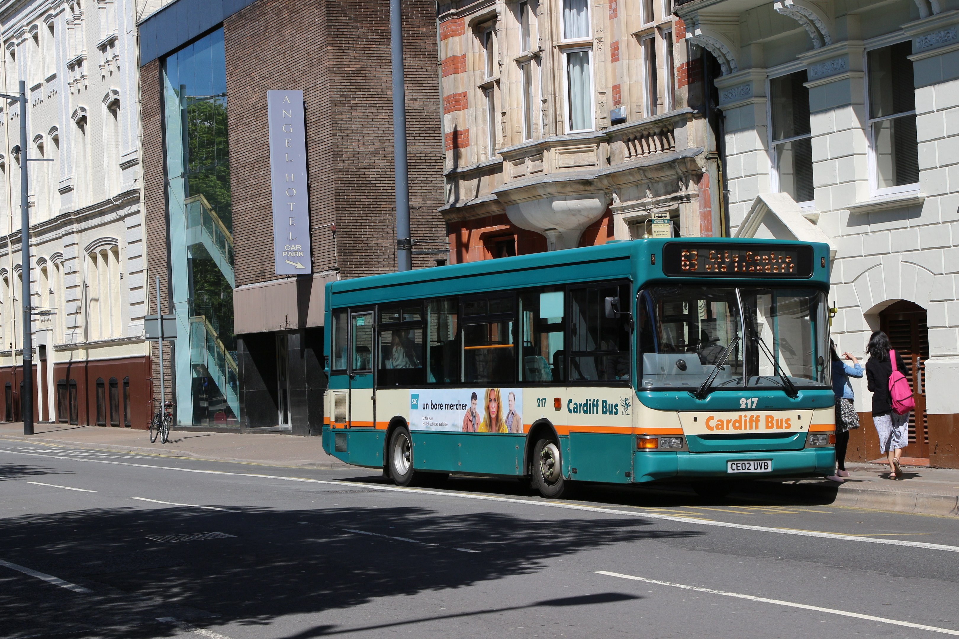 Ein blauer Bus fährt auf einer Straße mit hohen Gebäuden, während zwei Fußgänger auf dem Bürgersteig gehen, einer trägt eine Tasche, mit Strommasten, Schildern und einem Fahrrad im Hintergrund.
