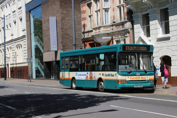 Ein blauer Bus fährt auf einer Straße mit hohen Gebäuden, während zwei Fußgänger auf dem Bürgersteig gehen, einer trägt eine Tasche, mit Strommasten, Schildern und einem Fahrrad im Hintergrund.