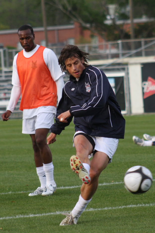 Zwei Fußballspieler, einer in schwarzer Jacke und weißen Shorts, die einen Ball kicken, der andere in orangefarbener Jacke dahinter, mit einem Zaun und Bäumen im Hintergrund.