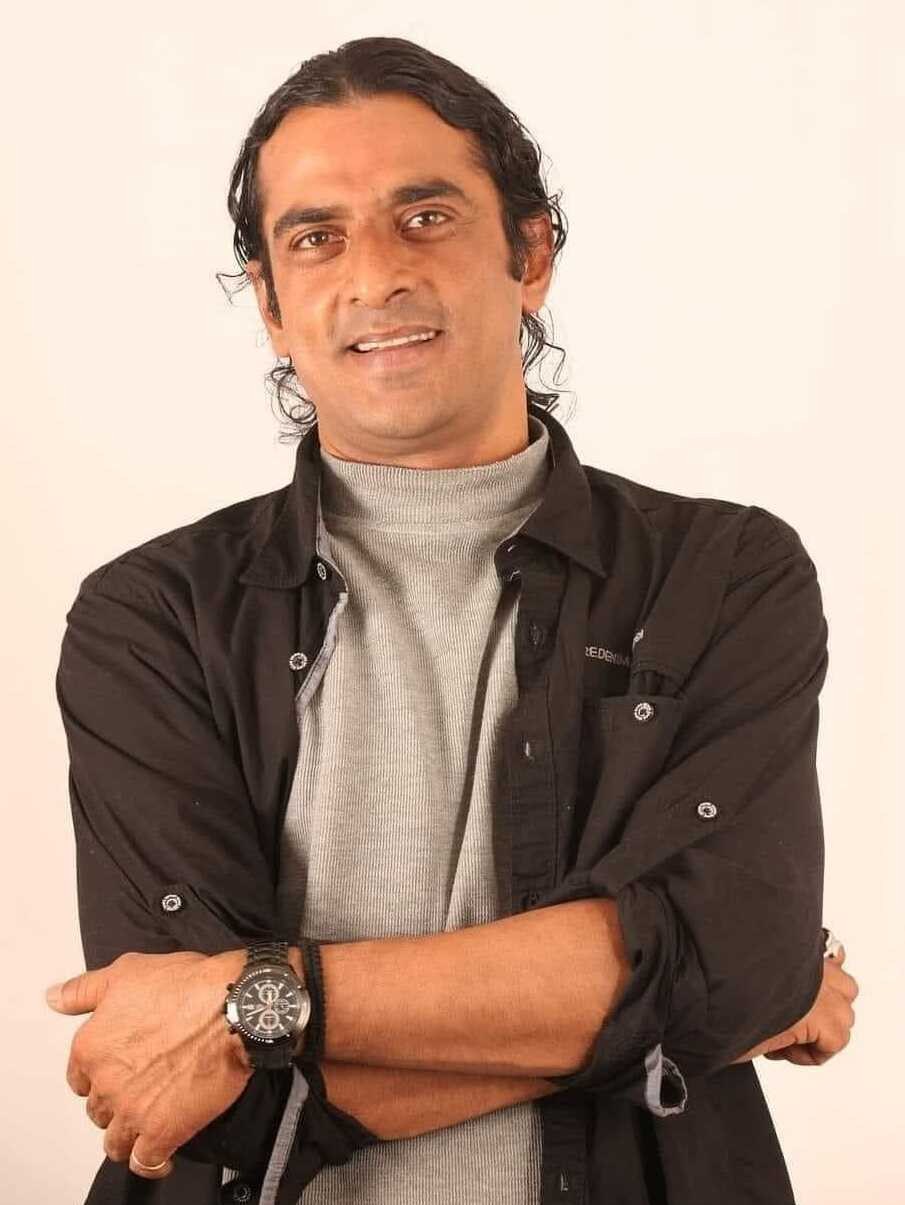 Ein Mann in einem schwarzen und aschfarbenen Kleid mit überschlagenen Armen und einer Uhr steht gegen einen weißen Hintergrund und sieht nachdenklich aus.