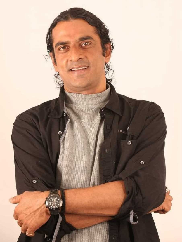 Ein Mann in einem schwarzen und aschfarbenen Kleid mit überschlagenen Armen und einer Uhr steht gegen einen weißen Hintergrund und sieht nachdenklich aus.