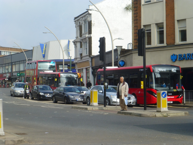 Ein roter Doppeldeckerbus fährt auf einer Stadtstraße mit hohen Gebäuden, Fußgängern auf dem Gehweg, einem Radfahrer, Verkehrsampeln, Laternen und Schildern unter einem bewölkten Himmel.