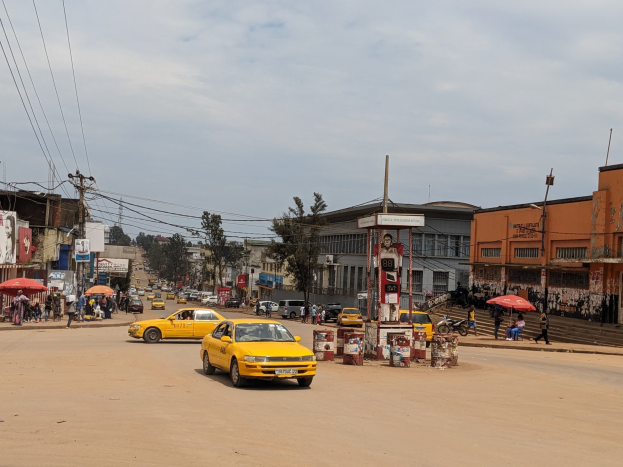 Ein gelbes Taxi fährt eine belebte Stadtstraße mit Gebäuden an den Seiten hinunter, während Fußgänger mit Regenschirmen auf dem Gehweg unter einem bewölkten Himmel gehen.