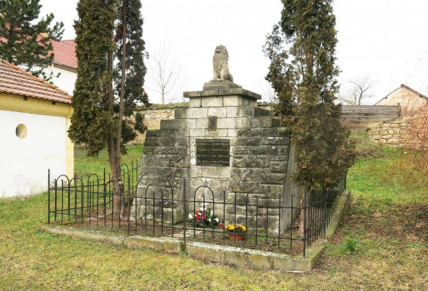 Steindenkmal für die Opfer des Holocaust in einer grünen Wiese, umgeben von einem Metallzaun, mit Blumensträußen drumherum, vor einer Kulisse aus Bäumen, Häusern, einer Wand und einem klaren blauen Himmel.