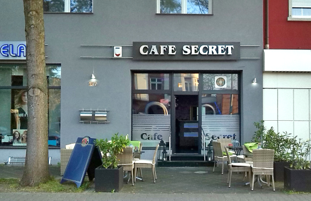 Ein Cafe in Berlin, Deutschland, mit einem Gebäude mit Fenstern und einer Tür, einem Schild mit Text, Außenbestuhlung mit Stühlen und Tischen, Topfpflanzen, einer Tafel, einem Baum und Gras.