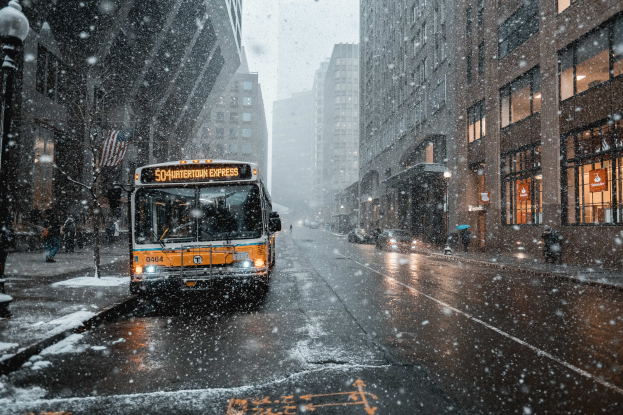 Ein gelber Bus fährt auf einer verschneiten Stadtstraße mit hohen Gebäuden auf beiden Seiten, Menschen auf dem Bürgersteig, Laternen und Bäumen, bei starkem Schneefall, der eine winterliche Atmosphäre schafft.