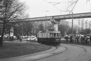 Ein Schwarz-Weiß-Foto einer Tram auf den Schienen, umgeben von Menschen, Fahrzeugen, einem Schild, einem Straßenpfahl, Bäumen, einer Brücke, Gebäuden mit Fenstern und einem bewölkten Himmel.