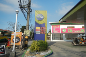 Tankstelle mit einem "Green's"-Schild, Fahrzeuge auf der Straße, umliegende Infrastruktur und Bäume unter einem bewölkten Himmel.
