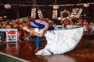 Eine Person küsst eine Frau in einer Außen-Szene mit Menschen an Tischen, Décoration, Bäumen und Lichtern im Hintergrund.