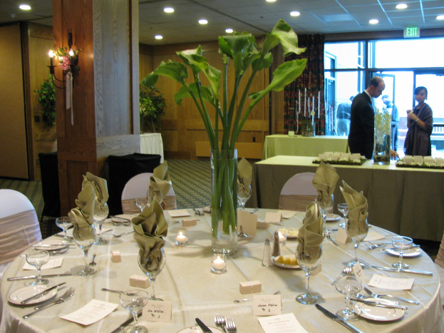 Ein Tisch mit einer wei├čen Decke, Tellern, Besteck, einem Glas und Zweigen, umgeben von St├╝hlen, mit einem gr├╝nen Tisch dahinter; ein Mann in einem schwarzen Blazer und ein M├Ądchen neben ihm, mit einer T├╝r dahinter und zwei M├Ąnnern dahinter.