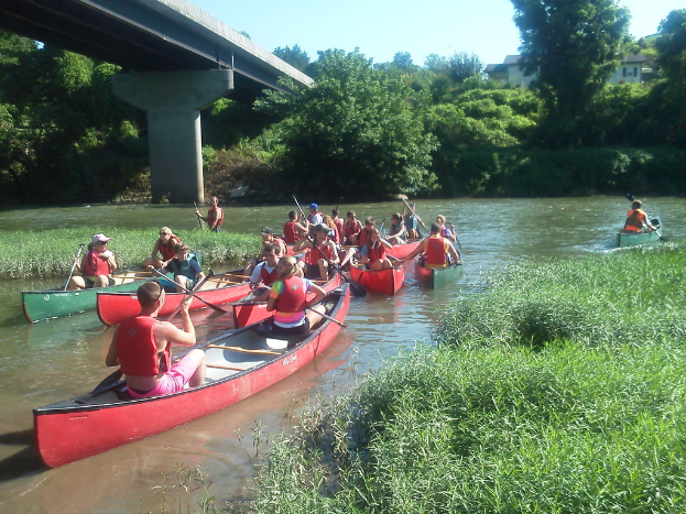 Eine Gruppe von Menschen auf Booten in einem See, mit Gras, einer Brücke, Bäumen, Pflanzen, Gebäuden und einem Himmel im Hintergrund.