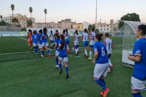 Eine Gruppe von Frauen, die Fußball auf einem Outdoor-Feld mit einem Torpfosten und Gebäuden im Hintergrund spielen.