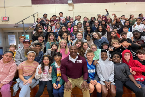 Gruppe von Schülern, die in einem Turnsaal für ein Foto posieren, wobei einige auf der Treppe sitzen und andere stehen, eine Tür und ein Ausgangsschild links sichtbar