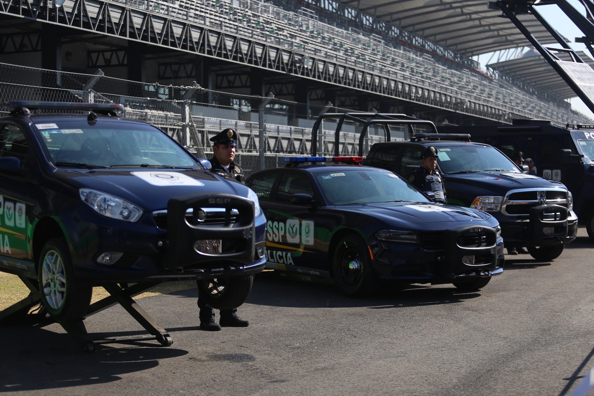 Eine Gruppe von Polizeiwagen vor einer Rennstrecke geparkt, mit uniformierten Personen auf der Straße, im Hintergrund ein Stadion und Maschendrahtzaun.
