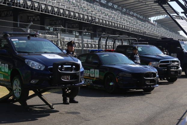 Eine Gruppe von Polizeiwagen vor einer Rennstrecke geparkt, mit uniformierten Personen auf der Straße, im Hintergrund ein Stadion und Maschendrahtzaun.