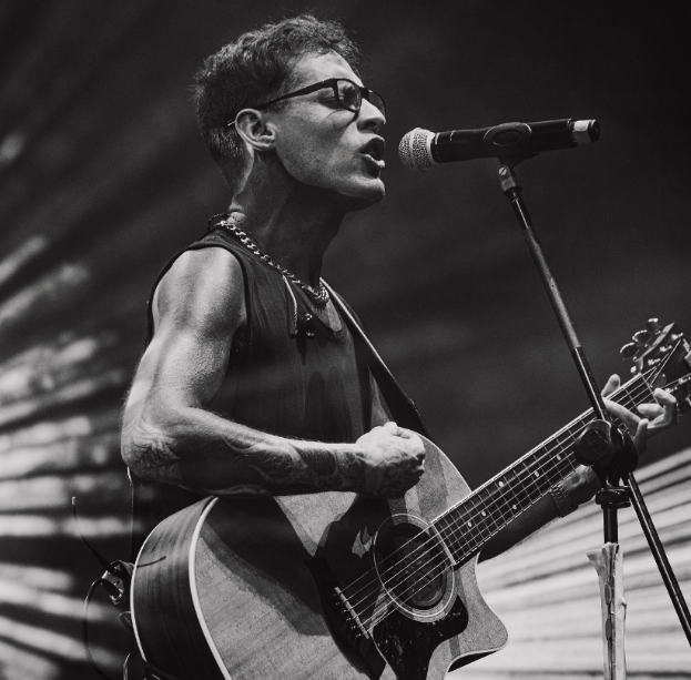 Ein Mann mit Brille spielt eine Gitarre vor einem Mikrofon und performt in der O2 Academy Brixton, dargestellt in Schwarz-Weiß.