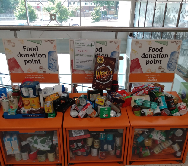 Orange Schübe mit Lebensmittelspenden, darunter Flaschen, Dosen und Kisten, mit Informationsschildern in der Nähe, durch Glasfenster betrachtet, die Außenfahrzeuge, Bäume, Gebäude und Himmel zeigen.