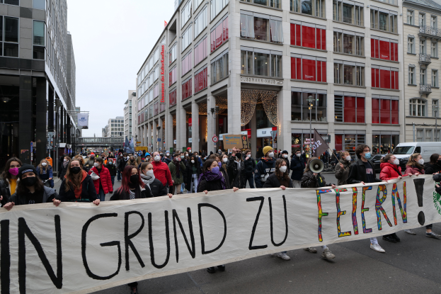 Eine Gruppe von Menschen mit Masken und einem Banner mit der Aufschrift "In Grund zu Feiern" steht vor Gebäuden mit Fenstern, Laternen und Fahrzeugen, was auf eine Demonstration in Berlin, Deutschland, hindeutet.
