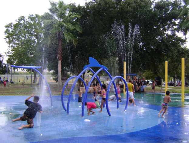 Ein Brunnen in der Mitte mit spielenden Kindern im Wasser, Menschen im Hintergrund sitzend und stehend, Büume und Fahrzeuge.