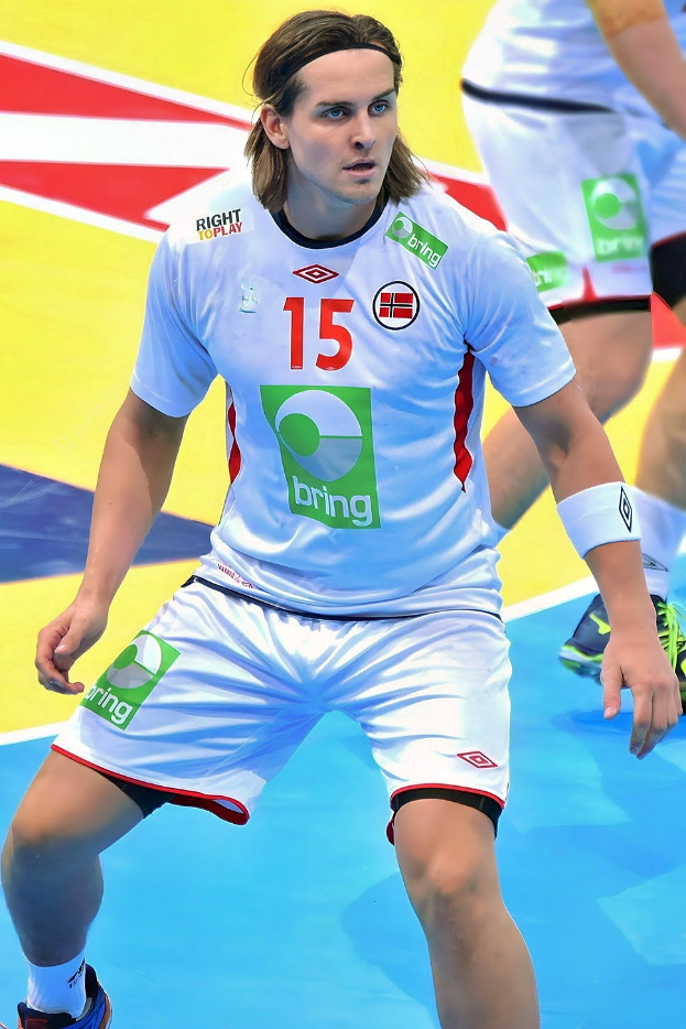 Ein Mann in weißer Uniform spielt Handball, mit zwei Personen im Hintergrund auf einem bunten Boden.