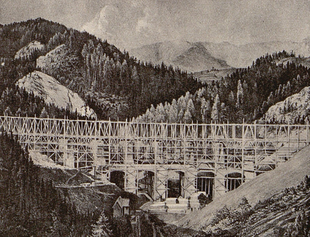 Ein Schwarz-Weiß-Foto einer Brücke im Bau in den Bergen, mit Bäumen, Hügeln und einem bewölkten Himmel im Hintergrund und einem Text am unteren Rand, der "Seilbrücke" lautet.