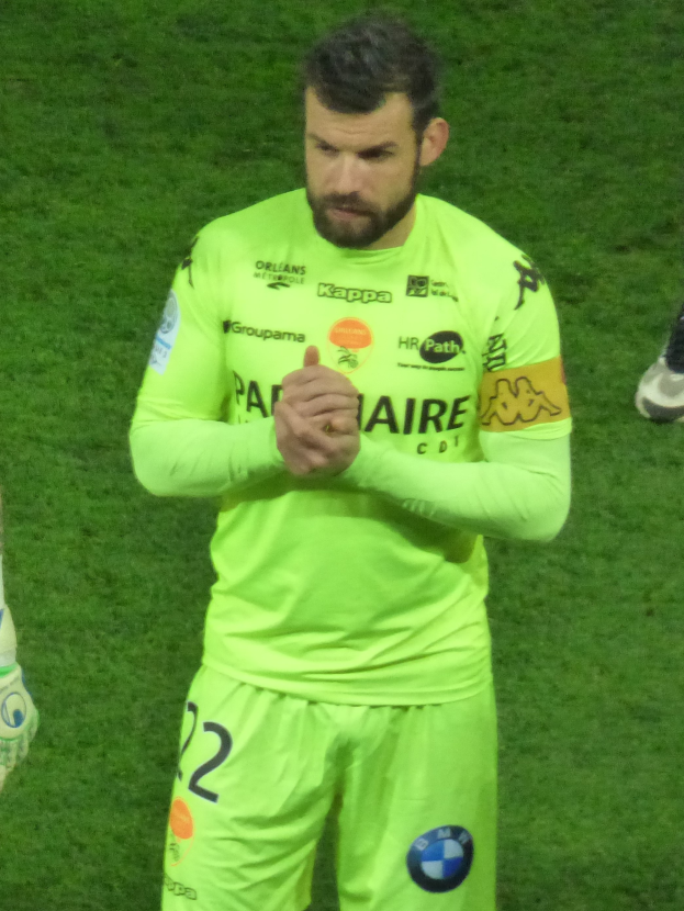 Ein Mann in einem gelben Torwarttrikot, der eine grüne T-Shirt und Shorts trägt, steht auf einem Fußballfeld und klatscht in die Hände, wobei ein anderer Personens Bein auf der linken Seite des Bildes zu sehen ist.