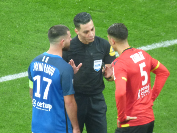 Schiedsrichter spricht mit zwei Männern auf einem Fußballfeld, einer trägt ein schwarzes Hemd und Hosen, der andere ein blau-rotes Hemd mit Text und Brille.