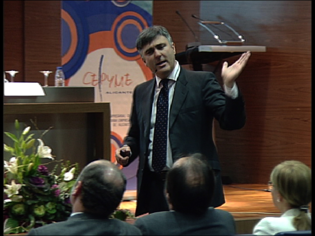 Ein Mann in einem schwarzen Blazer steht an einem Podium mit Mikrofonen, spricht in ein tragbares Mikrofon und hat eine Blumenanordnung und einen Tisch mit einem Buch und einer Wasserflasche in der Nähe, während drei Personen auf Stühlen vor ihm sitzen, vor einer braunen Wand und einem bunten Schild mit Text.
