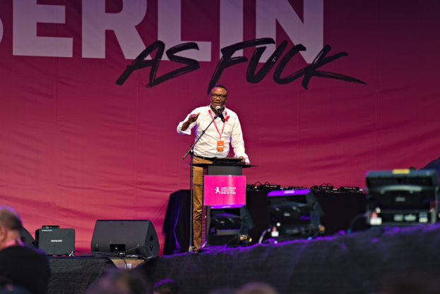 Mann in weißem Hemd und braunen Hosen, der in ein Mikrofon spricht, vor einem pinken 'Berlin as Fuck'-Plakat auf der Bühne.