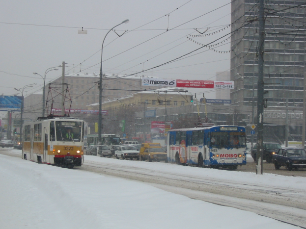 Eine städtische Straße mit viel Verkehr an einem verschneiten Tag, mit Fahrzeugen, Strommasten mit Drähten, Laternen, Gebäuden mit Fenstern und Schildern mit Text unter einem klaren Himmel.