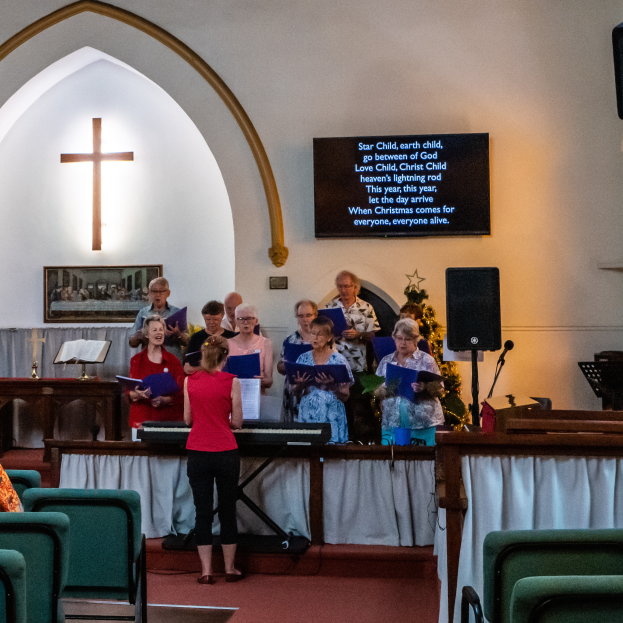 Eine Gruppe von Menschen steht vor einer Kirche, einige halten Bücher in den Händen, Stühle auf der linken Seite, ein Kreuzsymbol und ein Photo-Rahmen an der Wand, eine Tafel mit Text, ein Lautsprecher mit Ständer und ein Tisch mit einer Decke darauf, scheinbar singend als Chor.