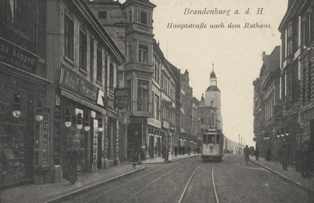 Ein altes Schwarz-Weiß-Foto einer Stadtstraße in Brandenburg, Deutschland, mit einer Tram auf Schienen, Menschen auf dem Gehweg, Gebäuden mit Fenstern, Schildern, Straßenlaternen und einem bewölkten Himmel.