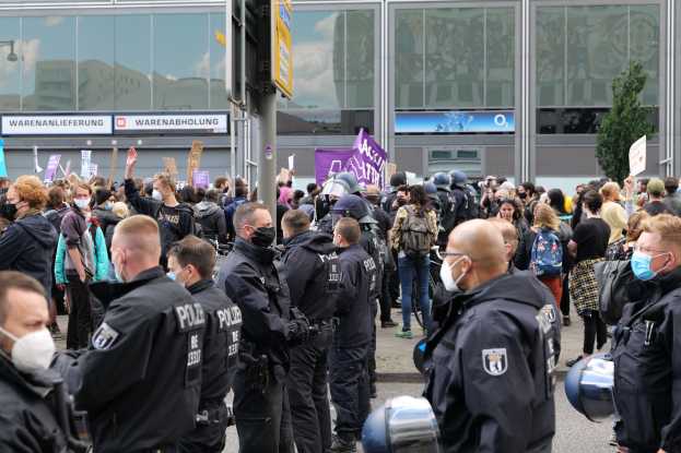 Große Gruppe von Menschen protestiert vor einem Gebäude, einige halten Schilder und tragen Helme, mit einem Schildständer und einem Baum im Vorder- und Hintergrund.
