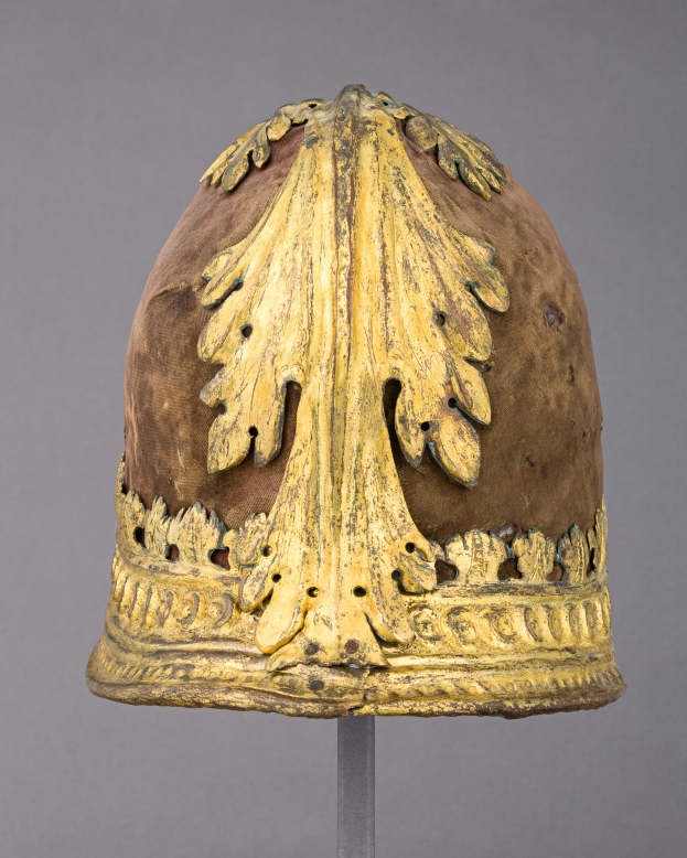 Gold-kronierter Helm mit detaillierten Verzierungen auf einem Ständer vor einem grauen Hintergrund.