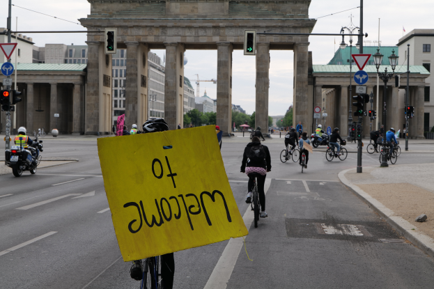 Eine Gruppe von Menschen auf Fahrrädern fährt eine Straße entlang vor dem Brandenburger Tor in Berlin, Deutschland, mit Helmen und einer Person, die ein gelbes Schild hält, mit Laternenpfählen, Verkehrszeichen, Gebäuden, Bäumen und einem klaren blauen Himmel im Hintergrund.