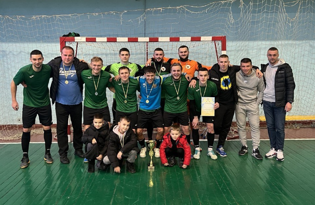 Eine Gruppe von Männern in Sportuniformen steht zusammen auf einem Futsalplatz, trägt Medaillen und hält einen Pokal, mit einem Torpfosten und einer Wand im Hintergrund.