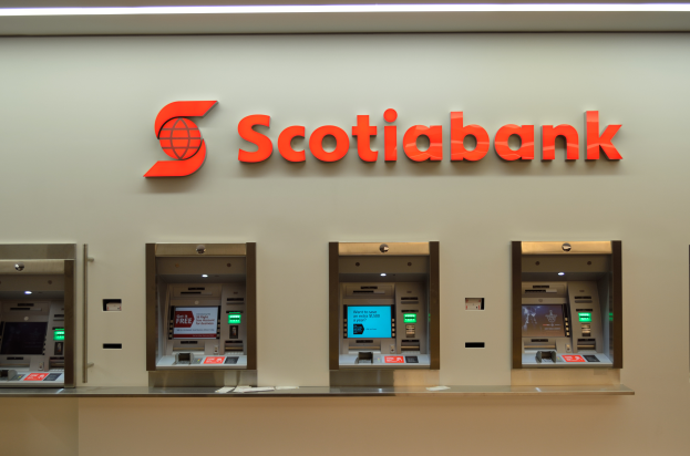 Ein modernes, elegantes Scotiabank-Geldautomatengerät in Schwarz und Silber mit einem großen Display und mehreren Knöpfen und Drehknöpfen, das an einer hellgrauen Wand mit dem Text "Schottische Bank" und einigen gerahmten Bildern hängt.