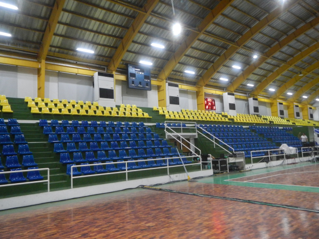 Großer Indoor-Sportplatz mit Holzboden, blauen und gelben Sitzplätzen, Geländern, Treppen mit Geländern, wandmontierten Bildschirmen, Deckenleuchten und einigen Personen.