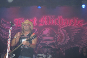 Eine Musik-Konzert-Szene mit Scheinwerfern oben, einem Banner im Hintergrund, Drogen in der Mitte unten, einer Person in der rechten unteren Ecke und einem Gitarristen mit Mikrofon links.