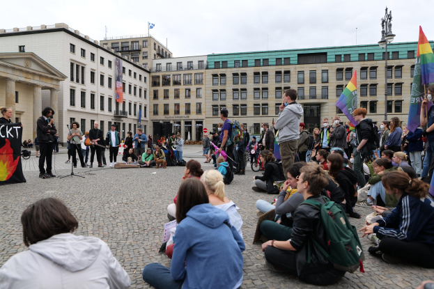 Eine Gruppe von Menschen, die auf dem Boden vor einer Menge sitzen, die Fahnen und Transparente hölt, während einer Demonstration gegen Homosexuelle in Berlin, mit einem Mikrofonständer, einer Statue auf einem Sockel und Gebäuden im Hintergrund bei einem bewölktem Himmel.