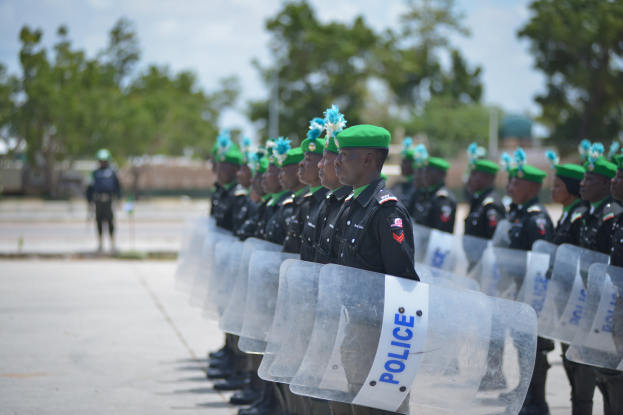 Eine Gruppe von Polizisten in schwarzen Uniformen und grünen Mützen steht in einer Reihe und hält durchsichtige Schilde, mit Bäumen, Gebäuden und einem klaren blauen Himmel im Hintergrund.